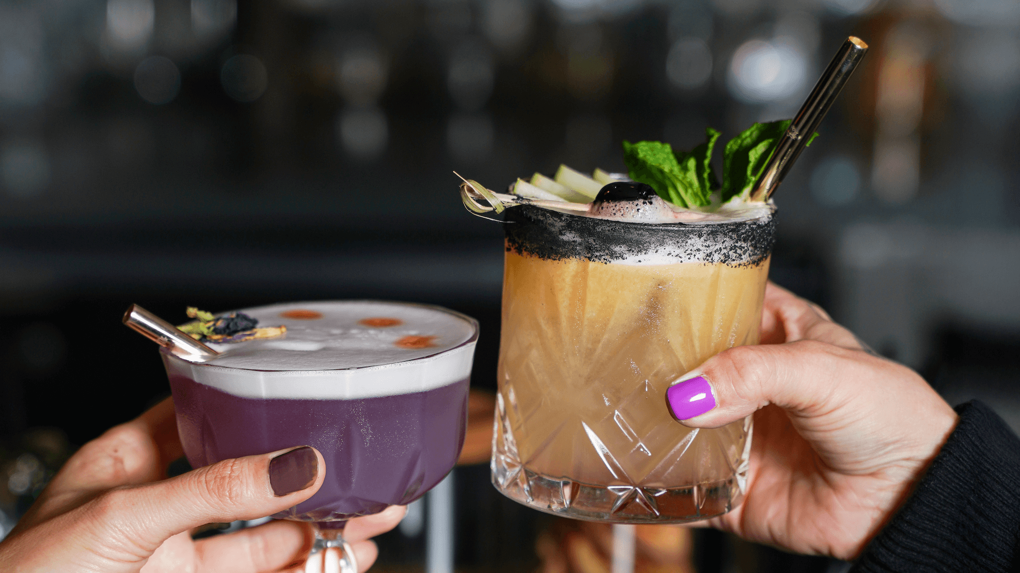 Two hands toasting with cocktails - one purple drink and one brown cocktail garnished with fresh mint, both featuring black reusable straws, showcasing Ecolux's eco-friendly luxury drinkware