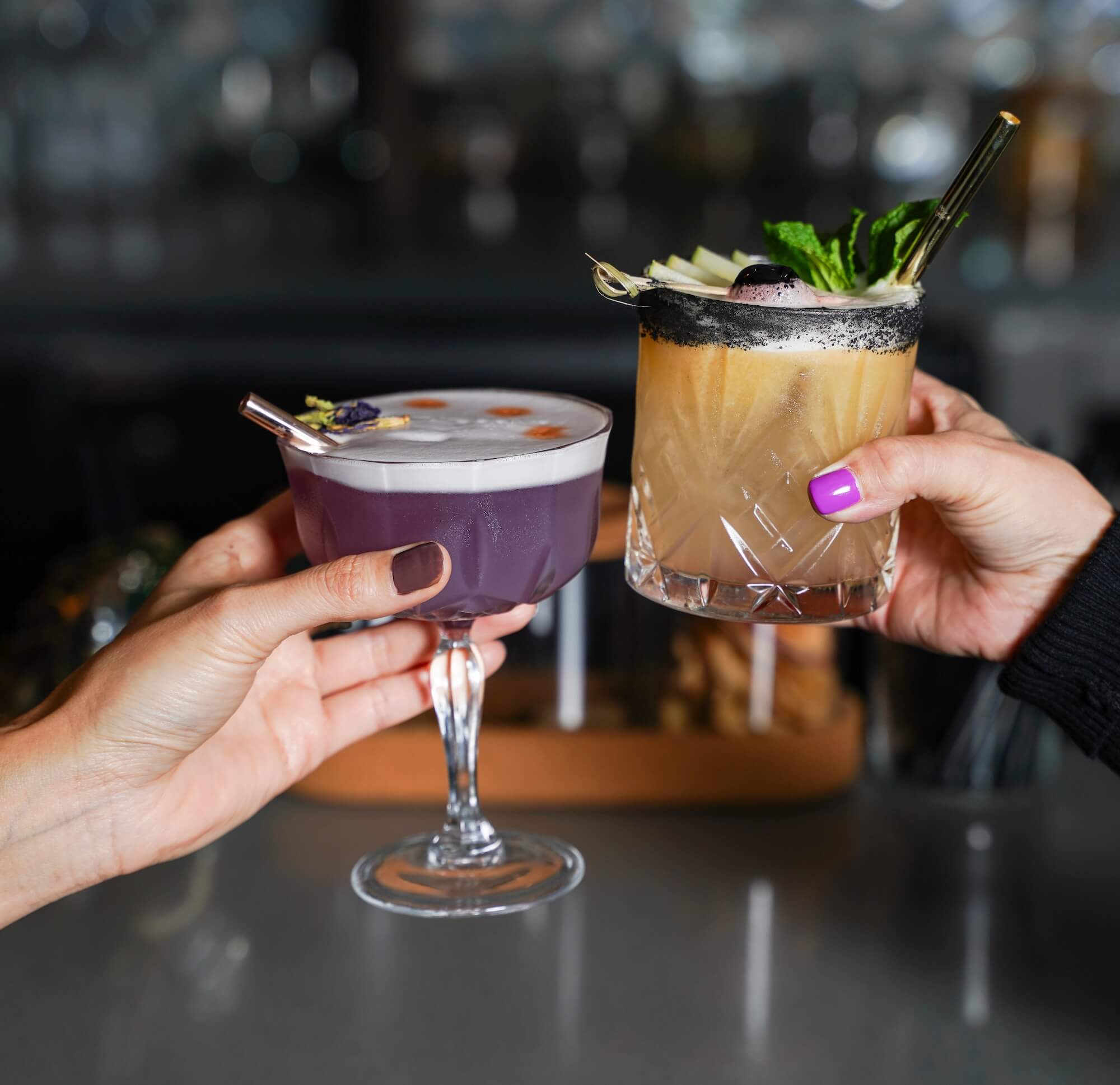 Two hands toasting with cocktails - one purple drink and one brown cocktail garnished with fresh mint, both featuring black reusable straws, showcasing Ecolux's eco-friendly luxury drinkware