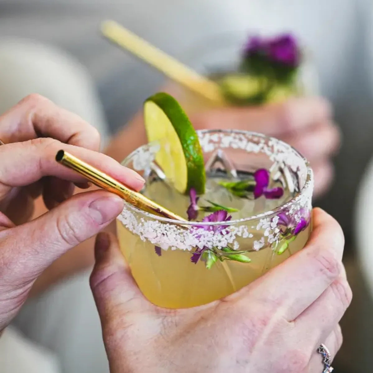 Hand holding margarita cocktail with salt rim, lime garnish, and edible purple flowers, using gold metal straw, with second cocktail visible in background