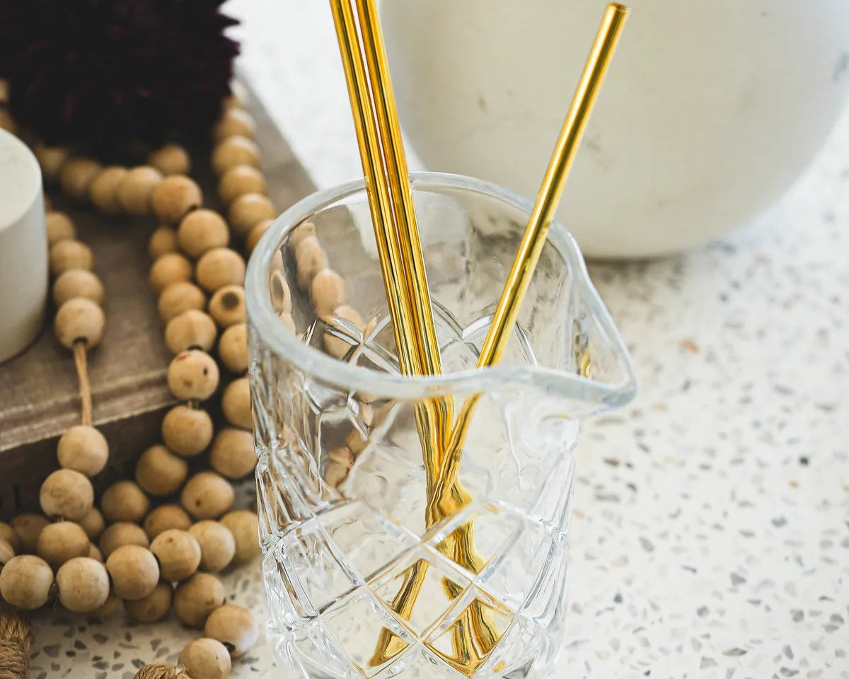 Multiple gold metal straws displayed in cut crystal glass on speckled countertop with decorative wooden beads and black flower in background