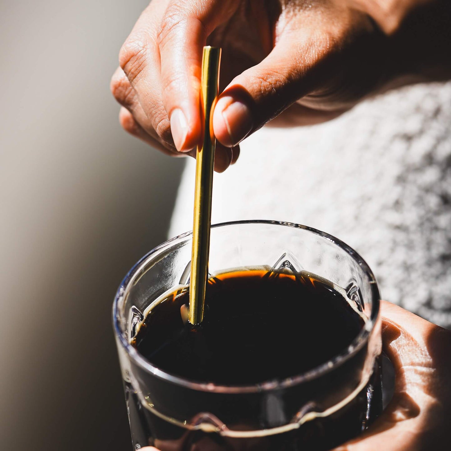 Person using gold metal straw to stir dark beverage in clear glass mug