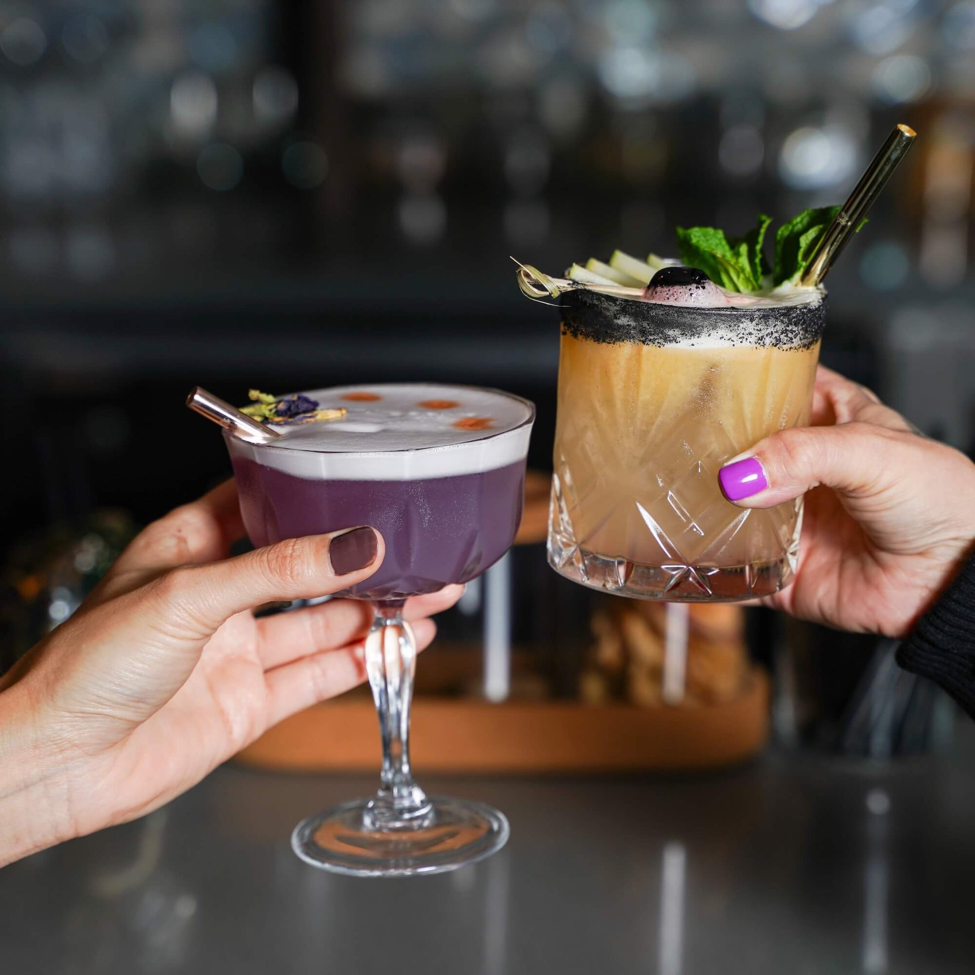 Dark cocktail in glass with rose gold metal straw and fresh herb garnish, person's hand visible